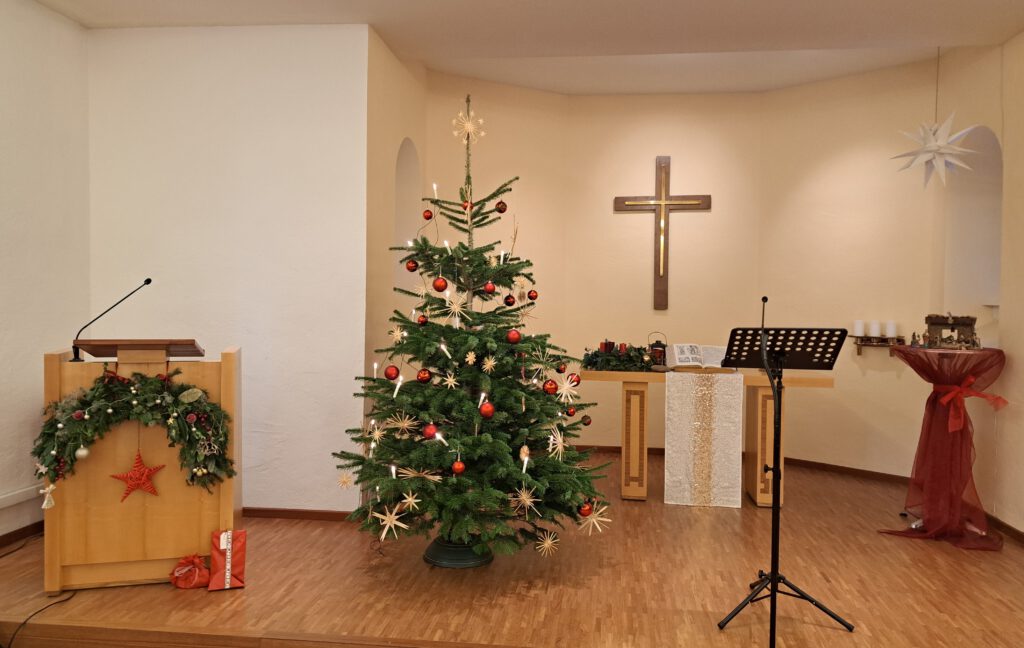 Blick in den weihnachtlich geschmückten Gottesdienstraum. Links sieht man eine Holzkanzel mit weihnachtlichem Halbkranz als Schmuck. Links vom Kreuz ein mit Strohsternen, roten Kugeln und Lichterkette geschmückten Weihnachtsbaum. Im Hintergrund in der Mitte Kreuz und Abendmahlstisch, rechts hängt ein weißer Herrnhuter Stern über einer kleinen Krippe auf einem Stehtisch.